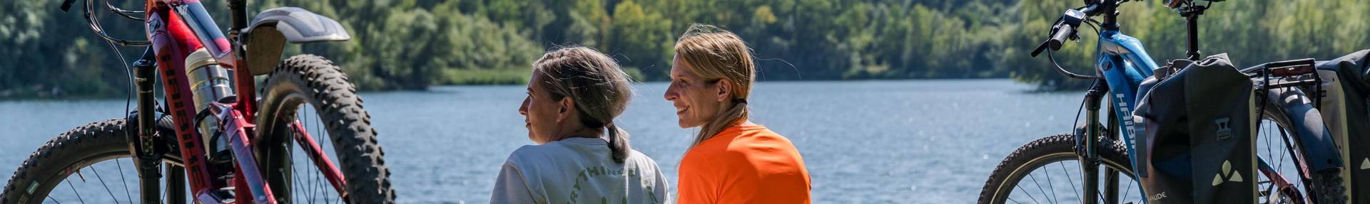 Zwei Frauen sitzen auf der Wiese am Ditfurter See und schauen auf den See. Links und rechts neben Ihnen steht ein Fahrrad.